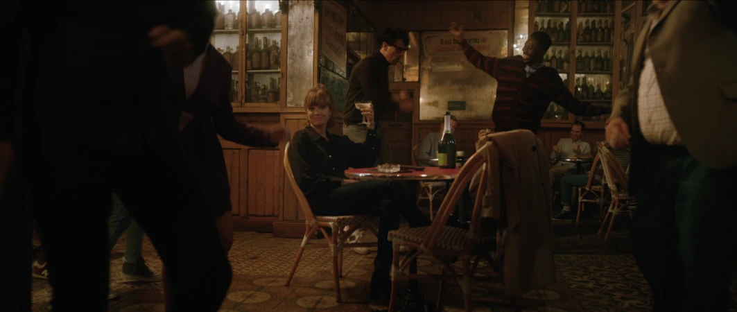 Woman sits with cocktail at dimly lit bar as others dance in the background. Wood paneling and patterned floor create an intimate atmosphere.