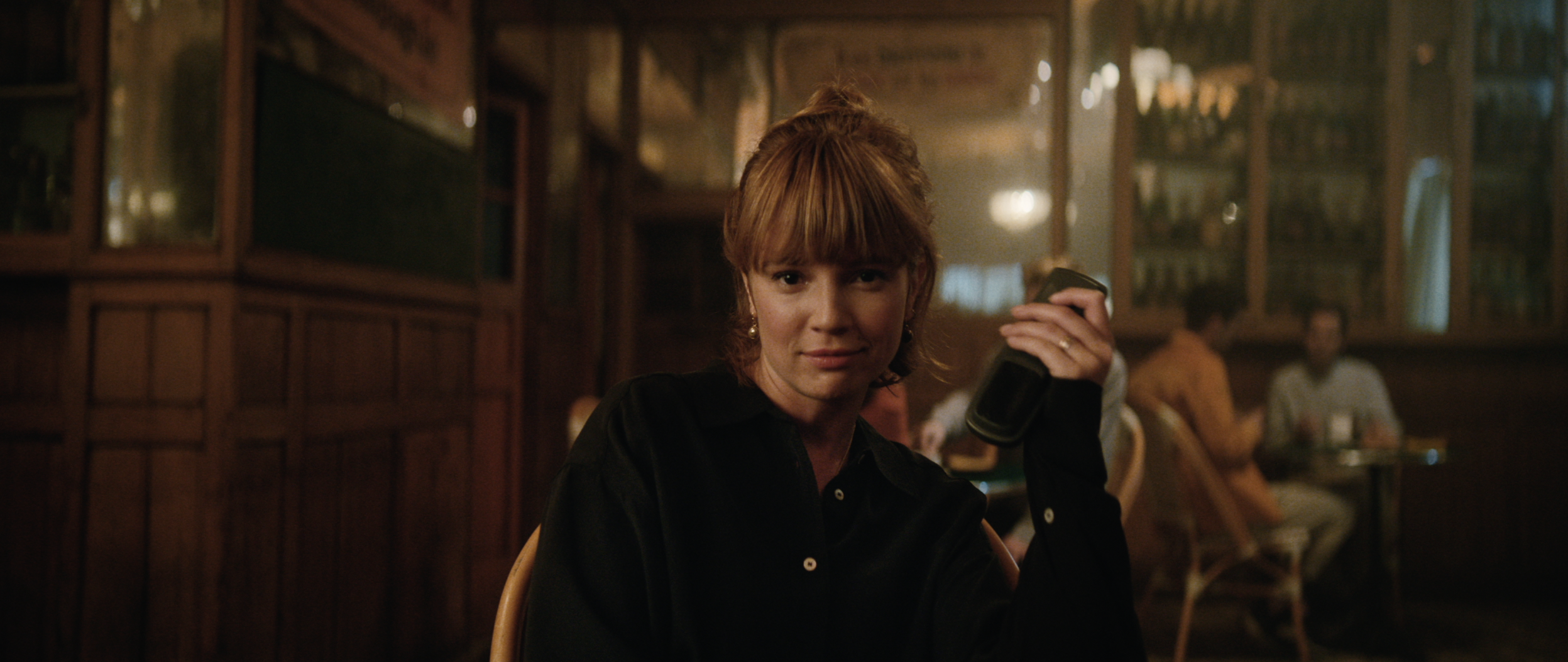 Attractive adult woman with auburn hair sits in an intimate bar setting, holding a black object, with patrons in the background.