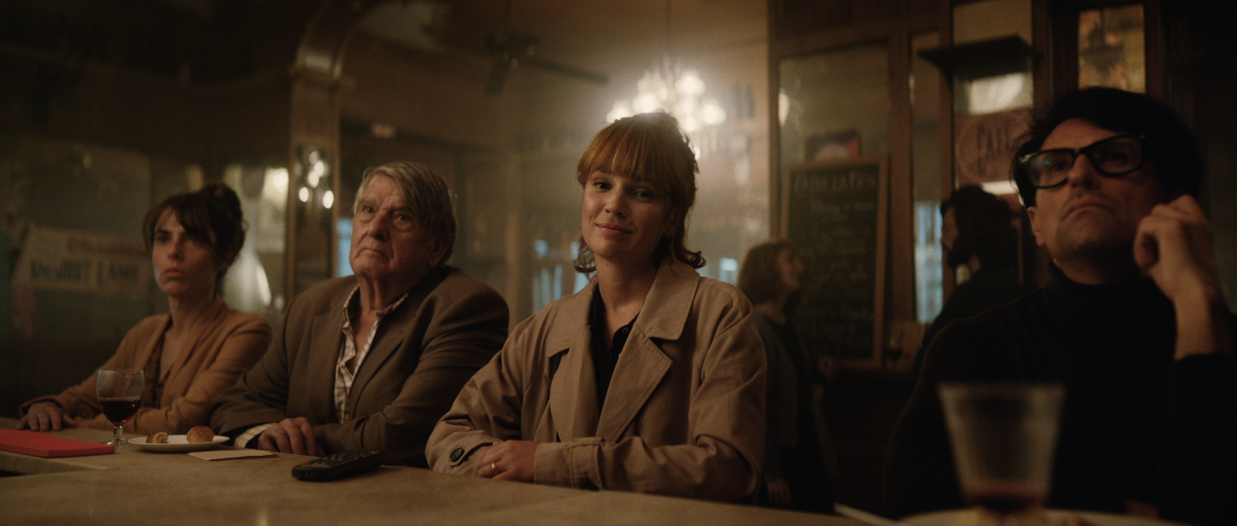 Four people sit at a bar with drinks and snacks. A woman in a beige coat smiles at the camera. Soft, warm lighting in the bar setting.