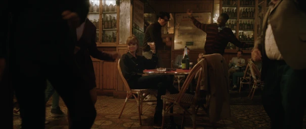 A woman seated at a bistro table holding a cocktail in a lively crowded bar directed by Nick Morissey