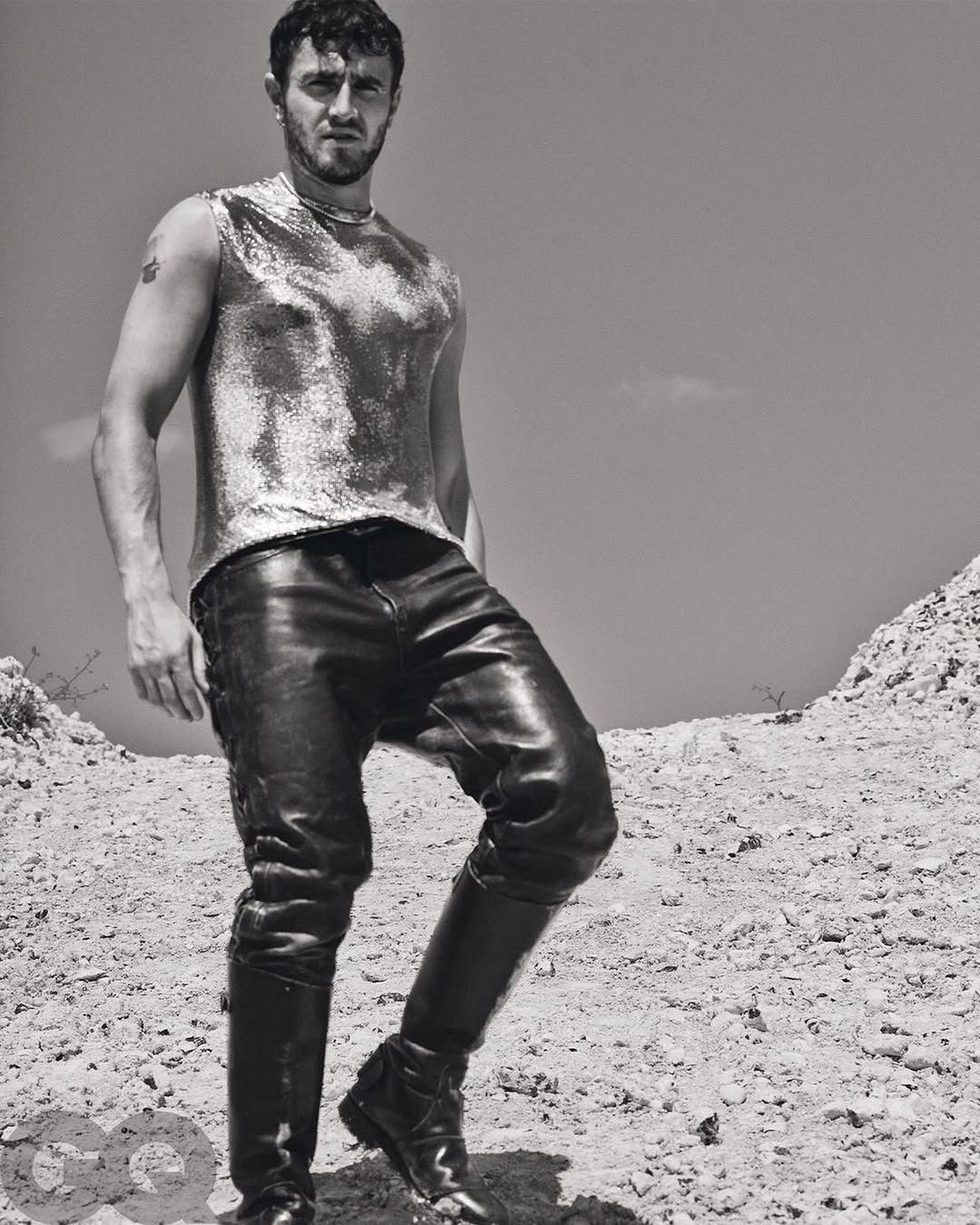 Monochrome portrait of Paul Mescal in metallic sleeveless shirt, leather trousers and boots, standing in a sparse, hilly landscape.