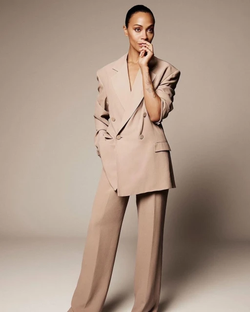 Portrait of a confident adult woman in a light brown double-breasted suit, striking a pose with hand near face against a neutral backdrop.