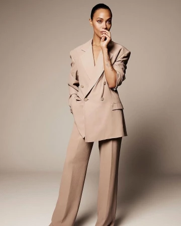 Portrait of a confident adult woman in a light brown double-breasted suit, striking a pose with hand near face against a neutral backdrop.