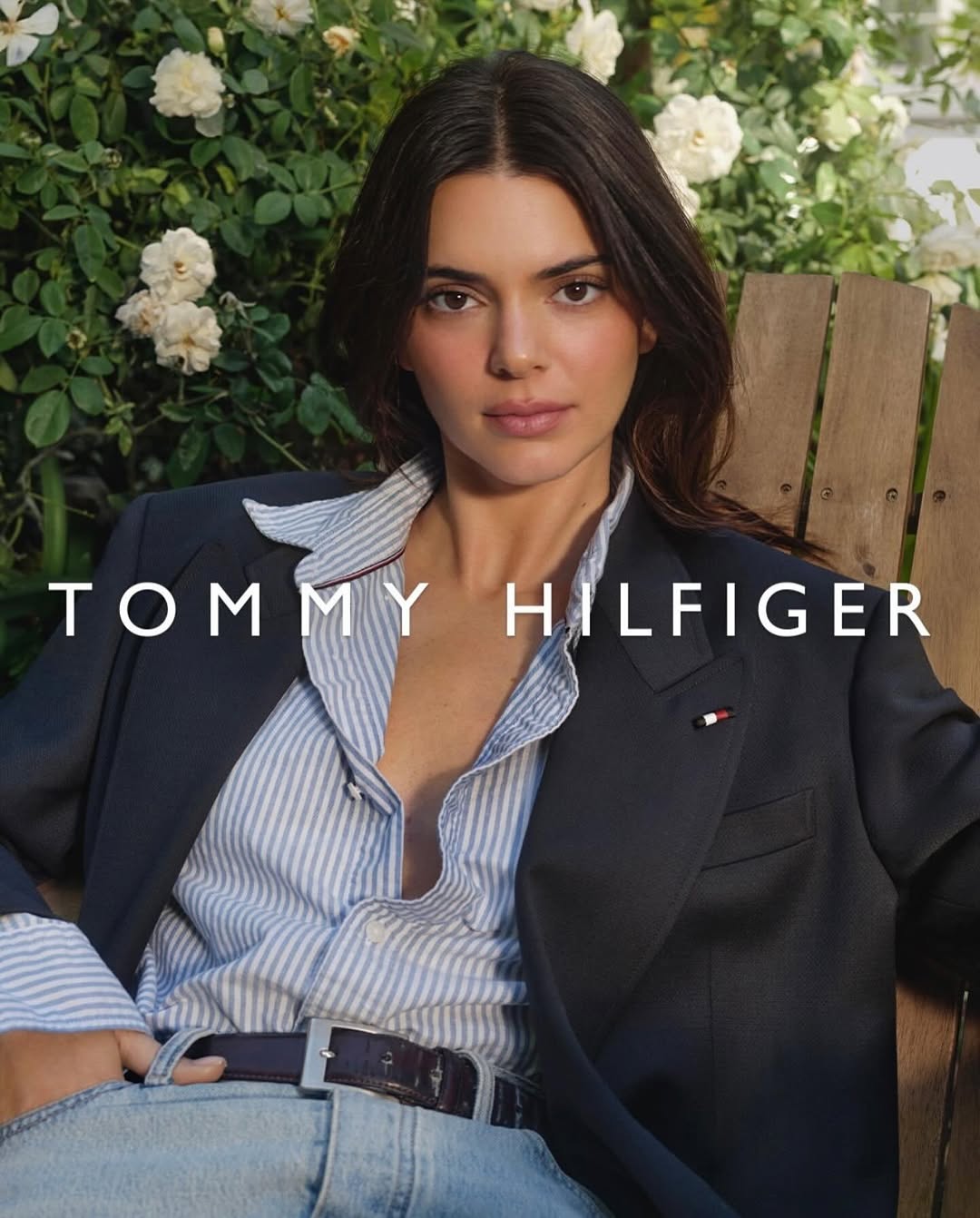 Portrait of a brunette woman wearing a navy blazer and striped shirt, sitting on a wooden bench with white roses in the background.
