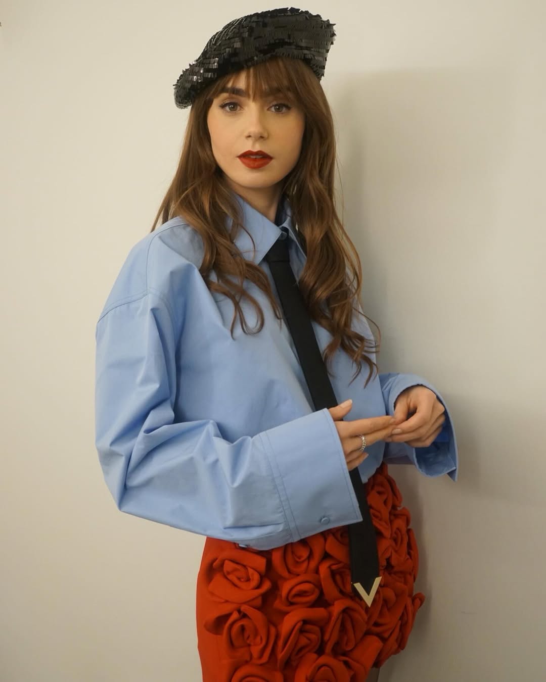 A woman with long brown hair and bangs poses indoors wearing a blue button-down shirt, black tie, red skirt, and black sequin beret.