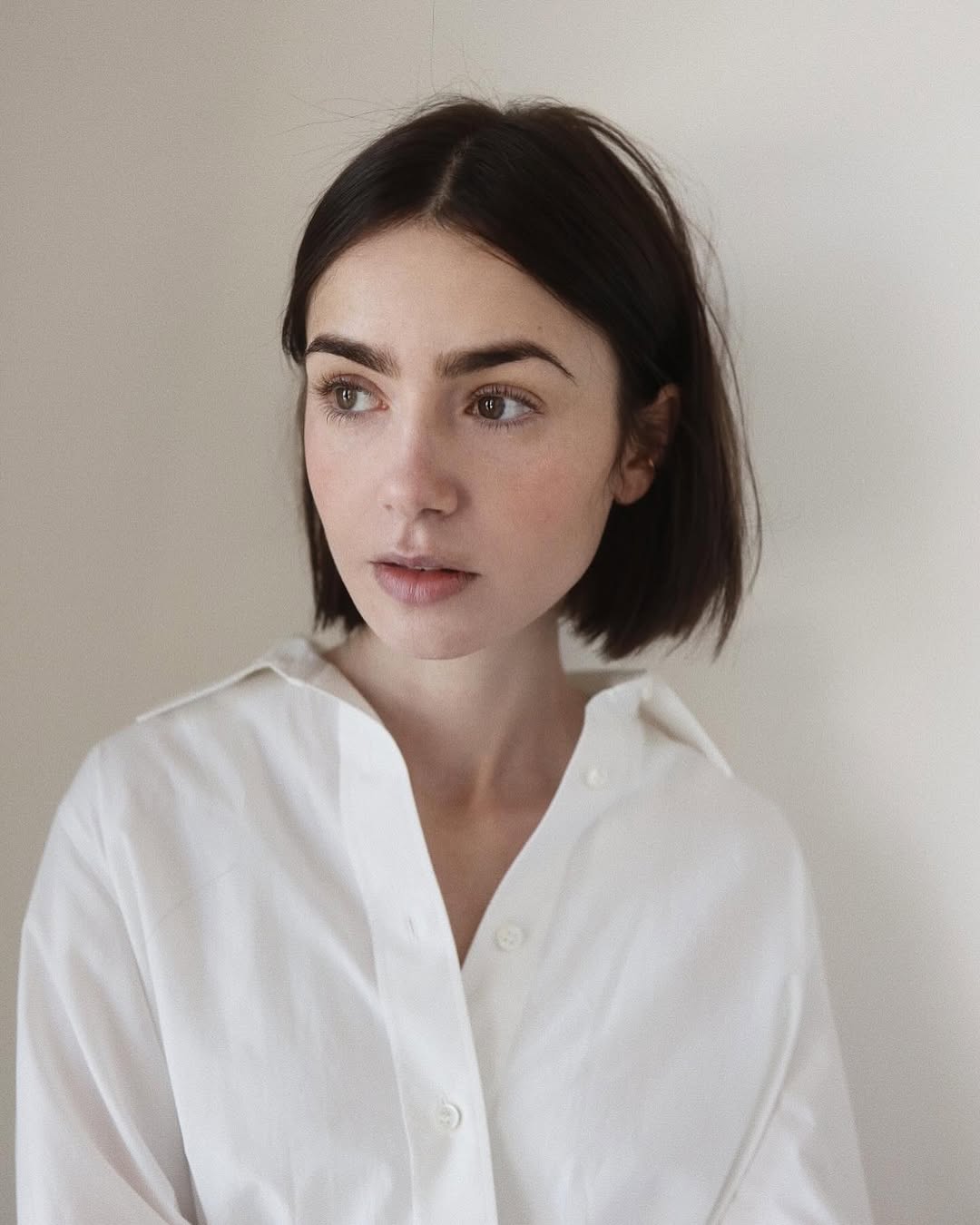 Portrait of a young woman with dark hair and a white button-down shirt, looking off to the side against a neutral background.
