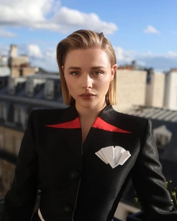 Portrait of blonde woman in black blazer with red accents and folded paper detail, posing against a blurred urban cityscape backdrop.