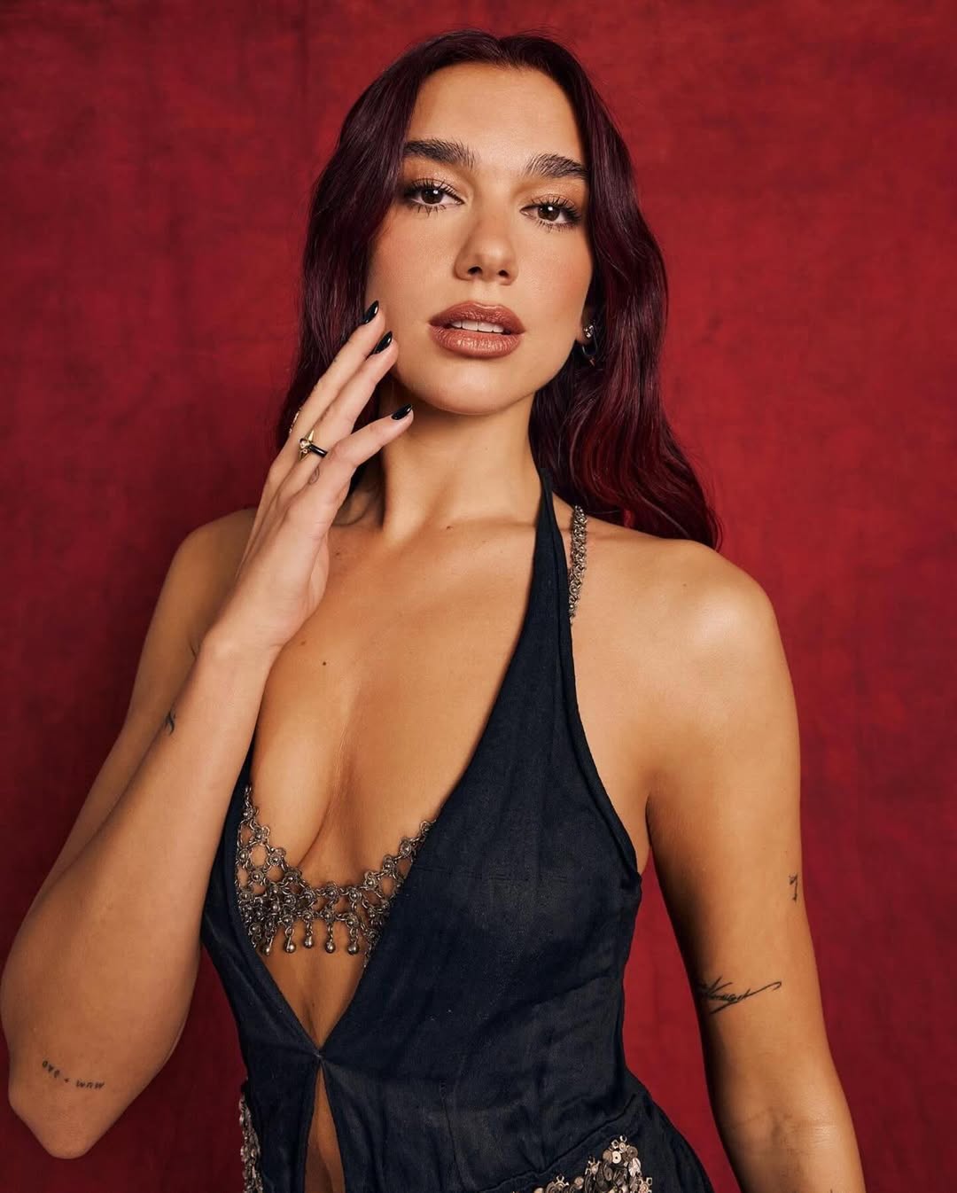 Portrait of a woman with dark hair in a black halter top, standing against a dark red background with dramatic lighting. 