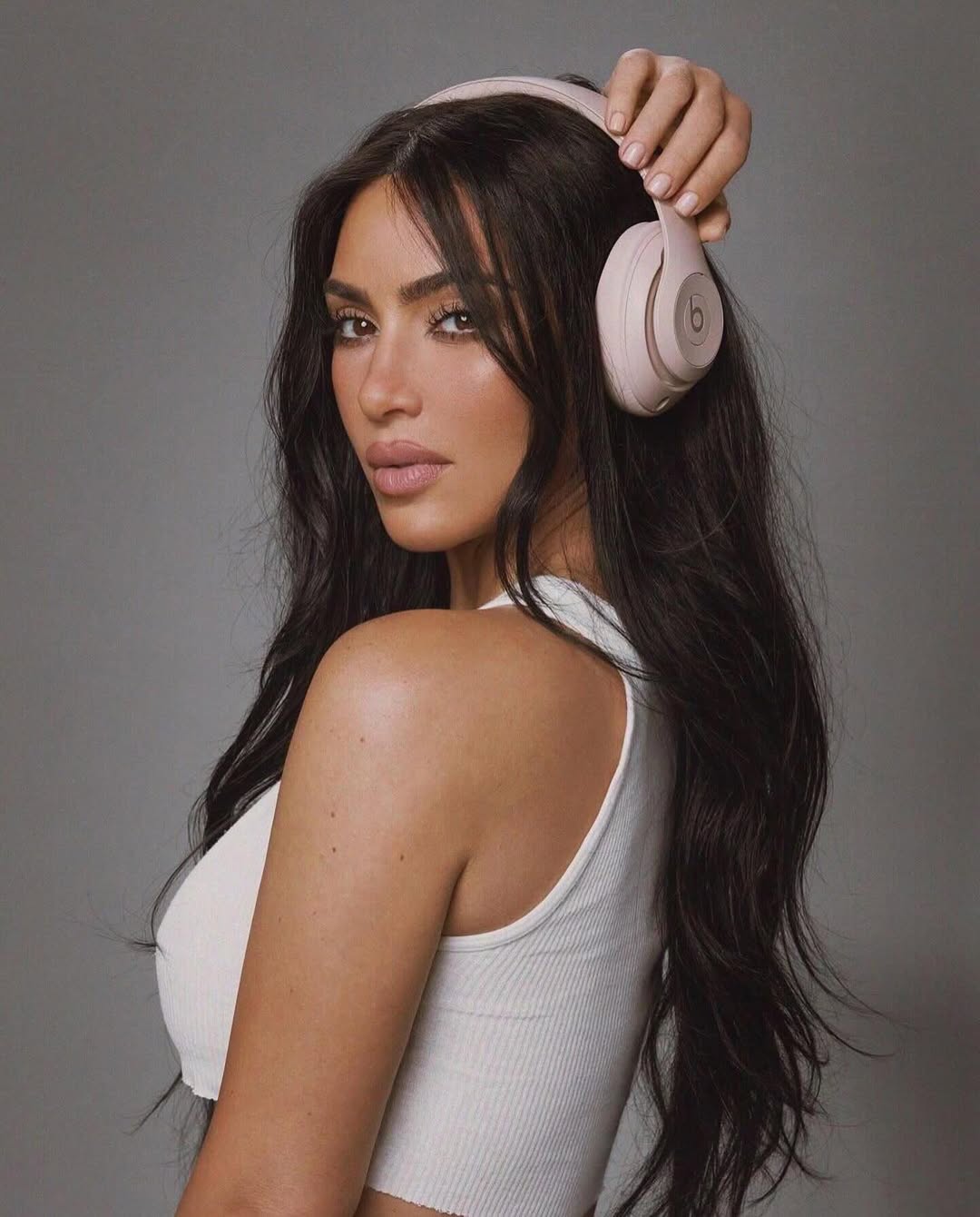 Portrait of a woman with long brunette hair holding pink headphones in a studio setting, wearing a white ribbed top, beauty shot.