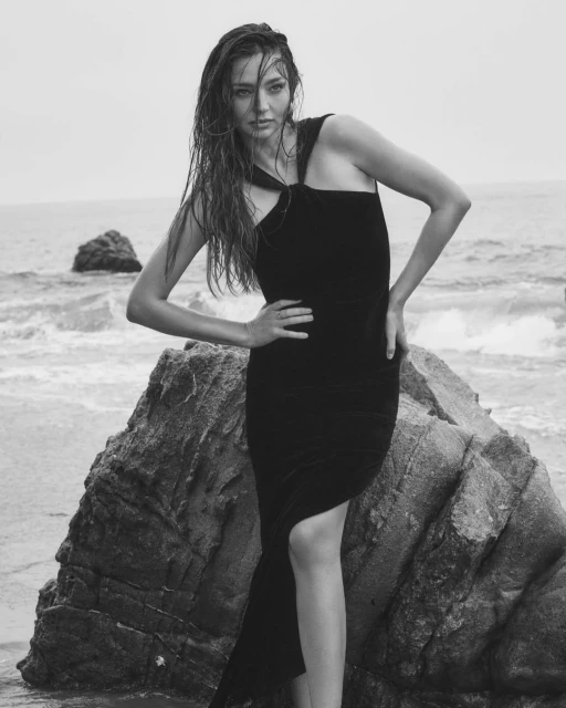 Monochrome image of a woman in a black dress posing on a rock at the beach. The ocean is in the background under an overcast sky.