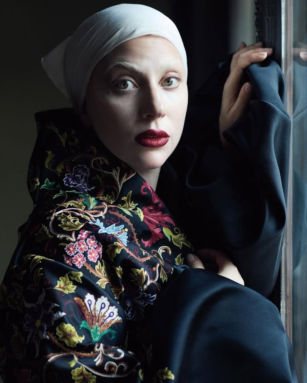 Close-up portrait of a woman with fair skin, white eyebrows, and red lipstick, wearing a white headscarf and an embroidered top.