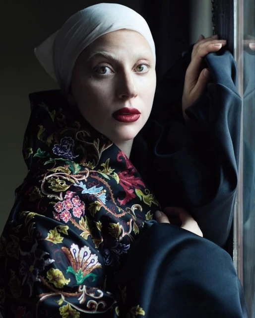 Close-up portrait of a woman with fair skin, white eyebrows, and red lipstick, wearing a white headscarf and an embroidered top.