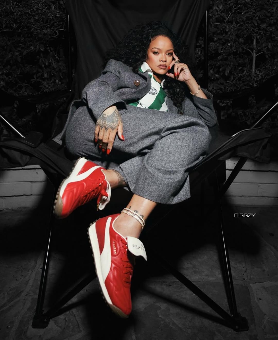Celebrity portrait of Rihanna sitting in a folding chair wearing a grey suit and red sneakers, posing outdoors against a dark backdrop.
