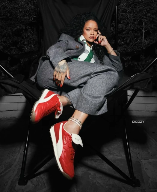Celebrity portrait of Rihanna sitting in a folding chair wearing a grey suit and red sneakers, posing outdoors against a dark backdrop.