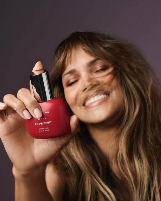 Adult female model smiles while holding a red bottle of JOYLUX LET'S SPIN hair product against a solid, dark purple background.