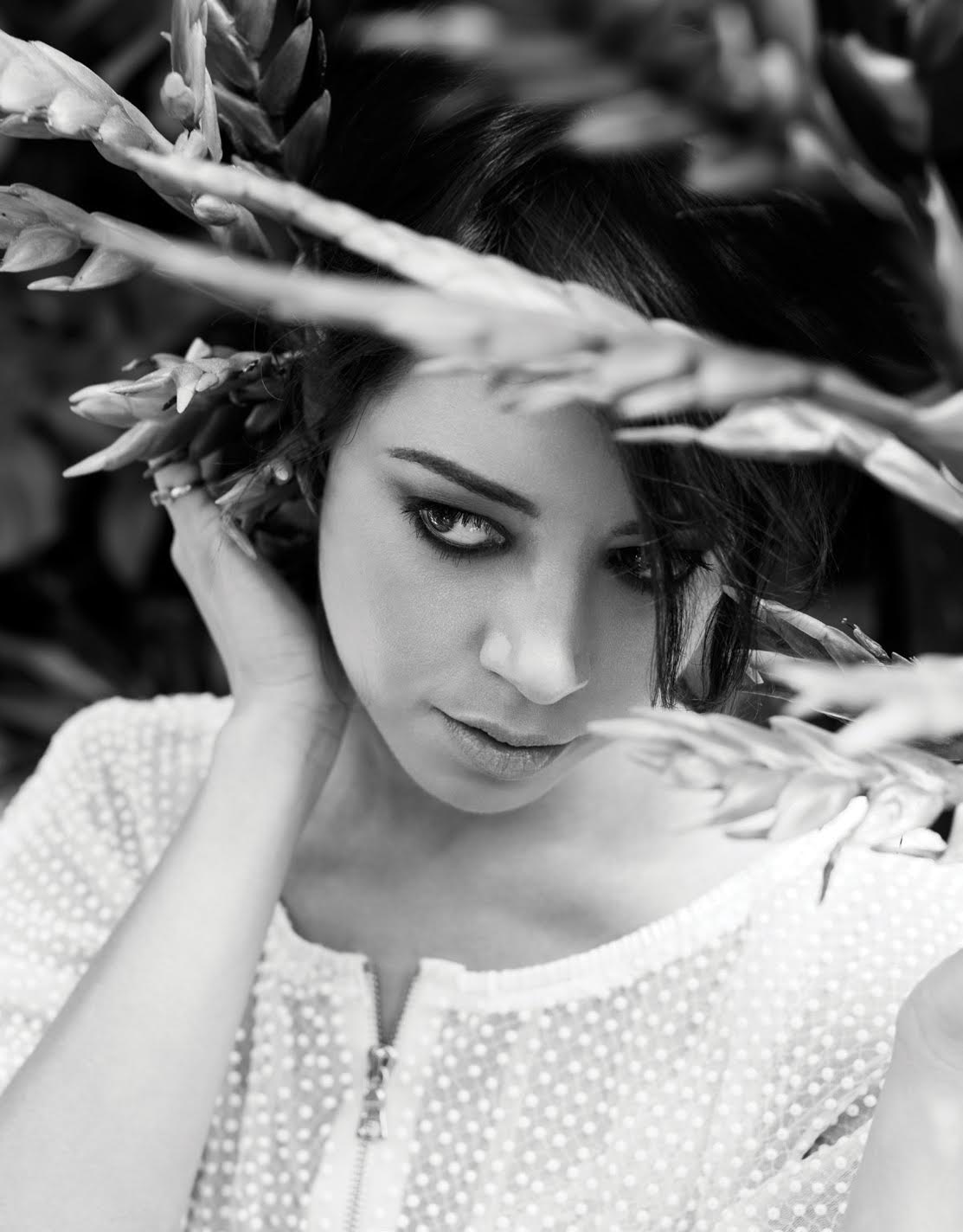 Portrait of a woman in black and white with dark hair, posed among tropical vegetation in an outdoor setting, wearing a textured shirt.