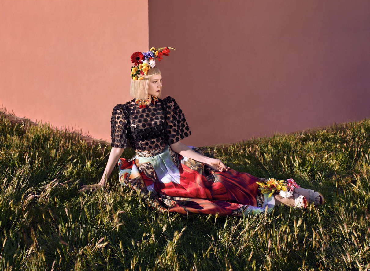 Woman in decorative floral headpiece and patterned dress sits in a grassy field with a peach-colored wall backdrop, editorial fashion.