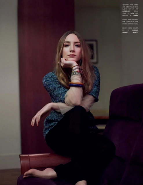 Woman with long blond hair in a teal sweater and black pants sitting in a purple velvet chair, looking at the camera.