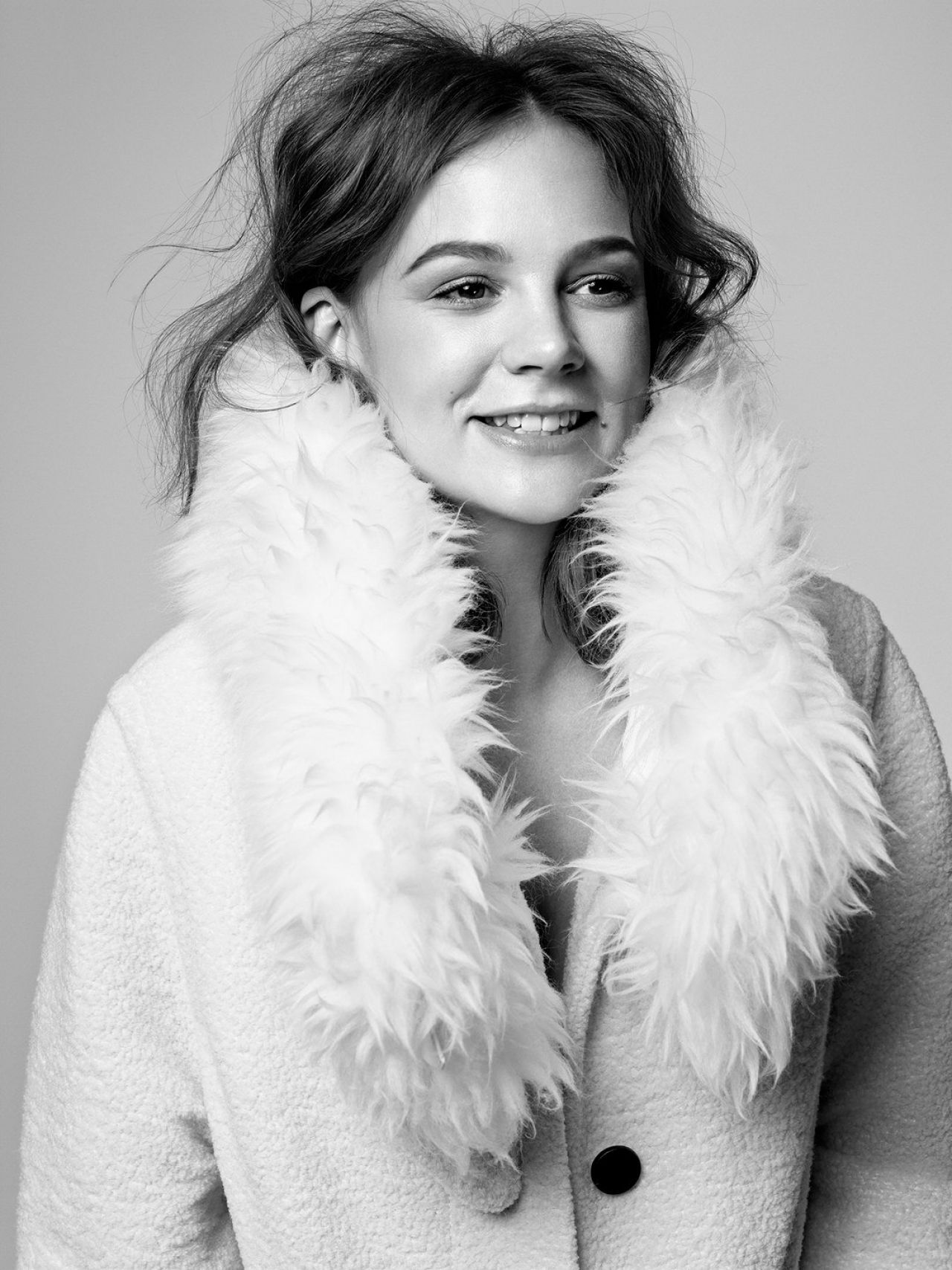Black and white portrait of a smiling woman wearing a fur-collared coat, looking to the side, with a plain background in a studio.