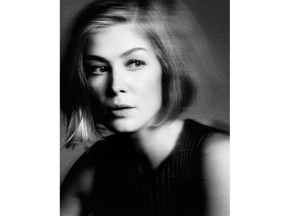 Black and white portrait of a woman with short blonde hair, wearing a dark top, posing against a blurred neutral background.