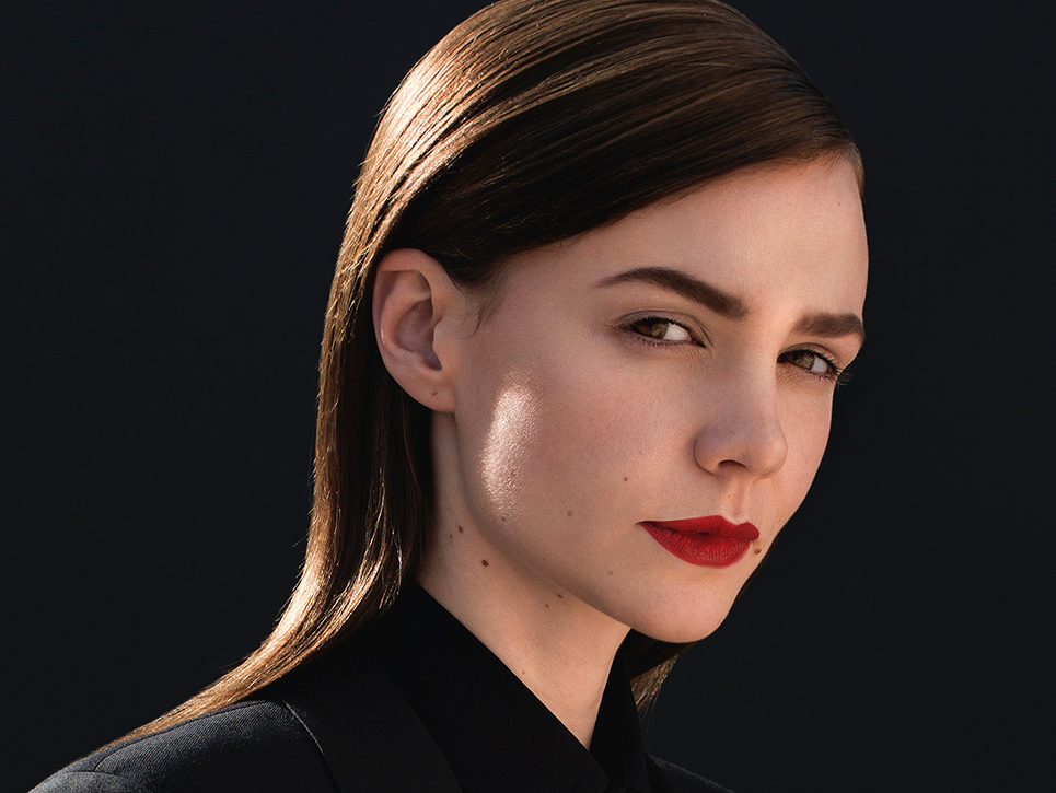 Close-up portrait of a woman with red lipstick, wearing a black jacket, against a dark background. Beauty shot with centered composition.