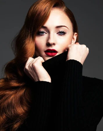 Portrait of Sophie Turner with long red hair and bold red lips pulling up a black turtleneck against a dark background