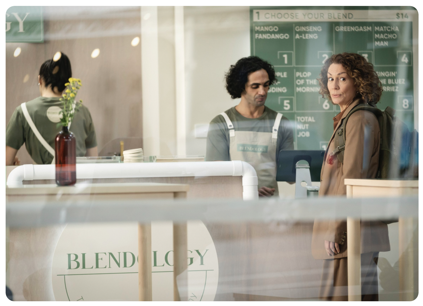 Woman with a backpack stands in line at a smoothie bar with a menu offering various blends as another employee works behind the counter.