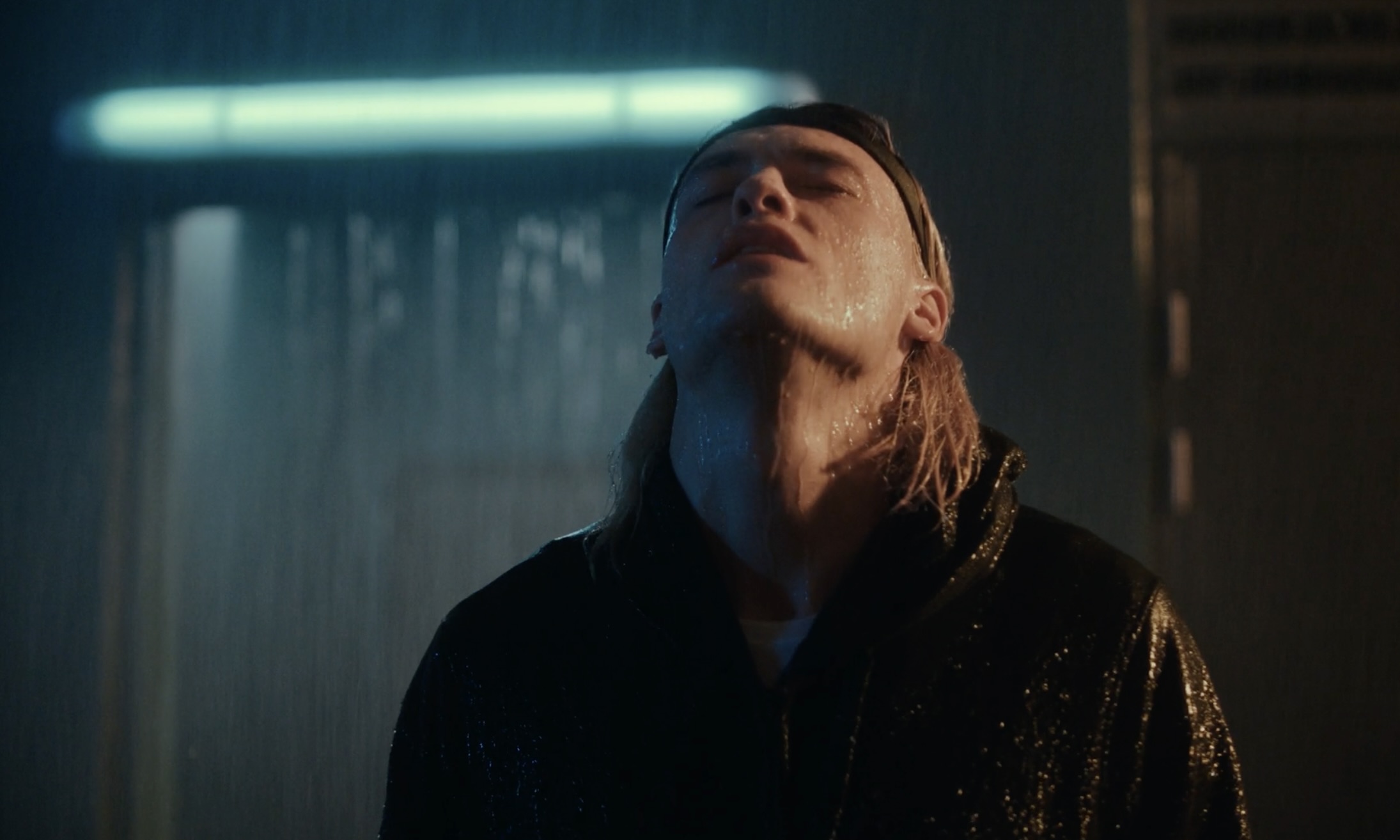Man in a black jacket stands in the rain with his head tilted back and mouth slightly open in an indoor environment.