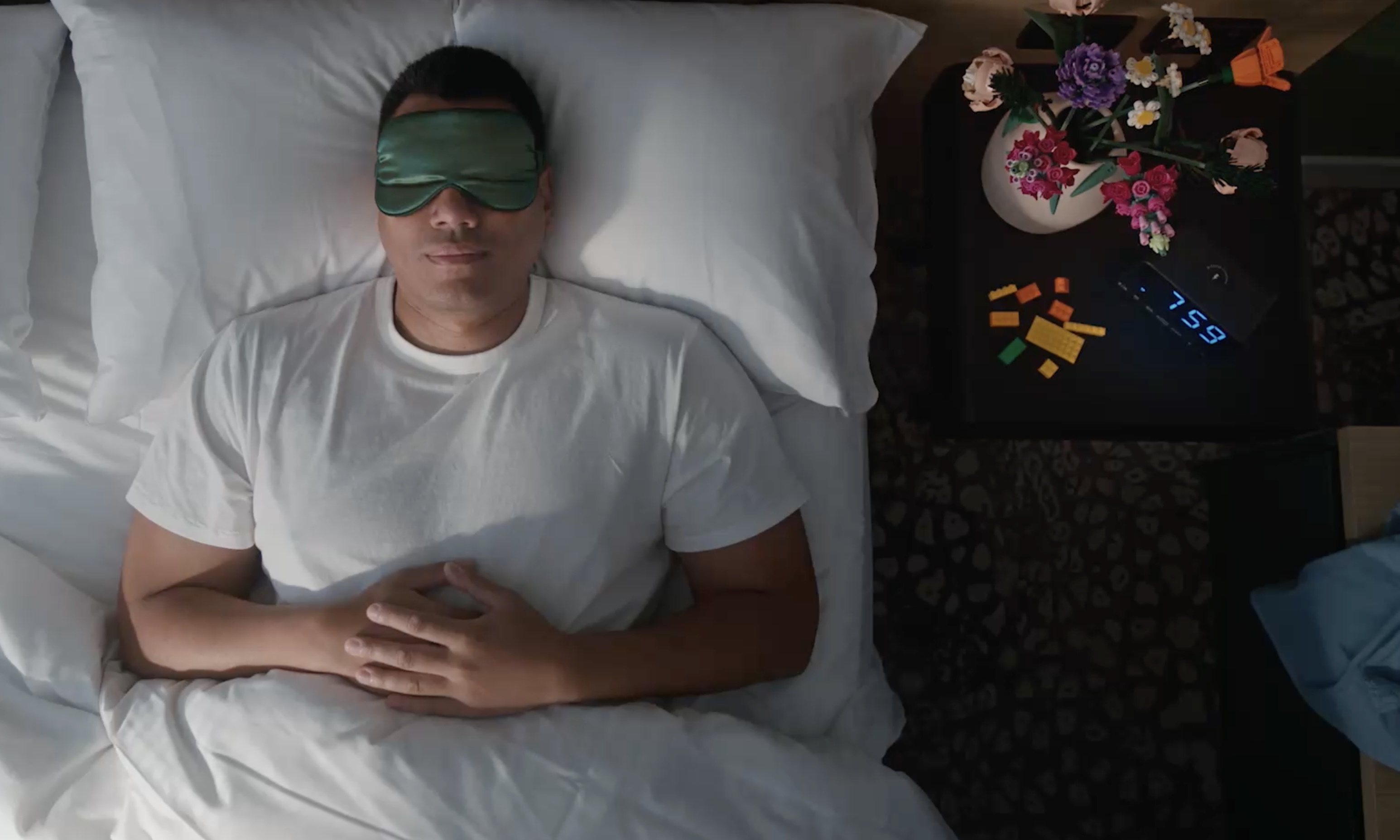 Adult male sleeping on white sheets with a green sleep mask, bedroom interior with a bedside table, lamp, alarm clock, and vase of flowers.