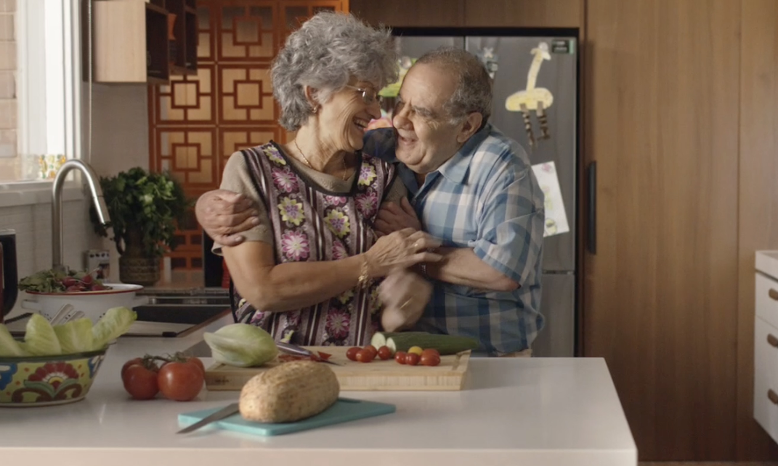 An elderly couple embraces in a bright, modern kitchen setting. Fresh produce, wooden cabinets, and stainless steel appliances visible.