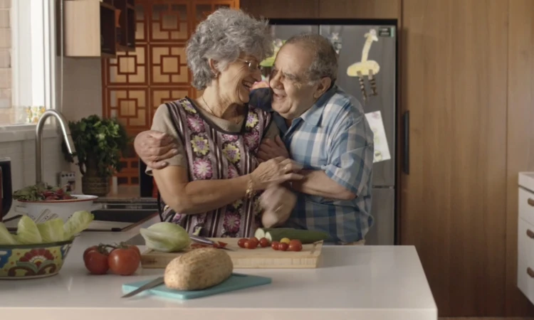 An elderly couple embraces in a bright, modern kitchen setting. Fresh produce, wooden cabinets, and stainless steel appliances visible.