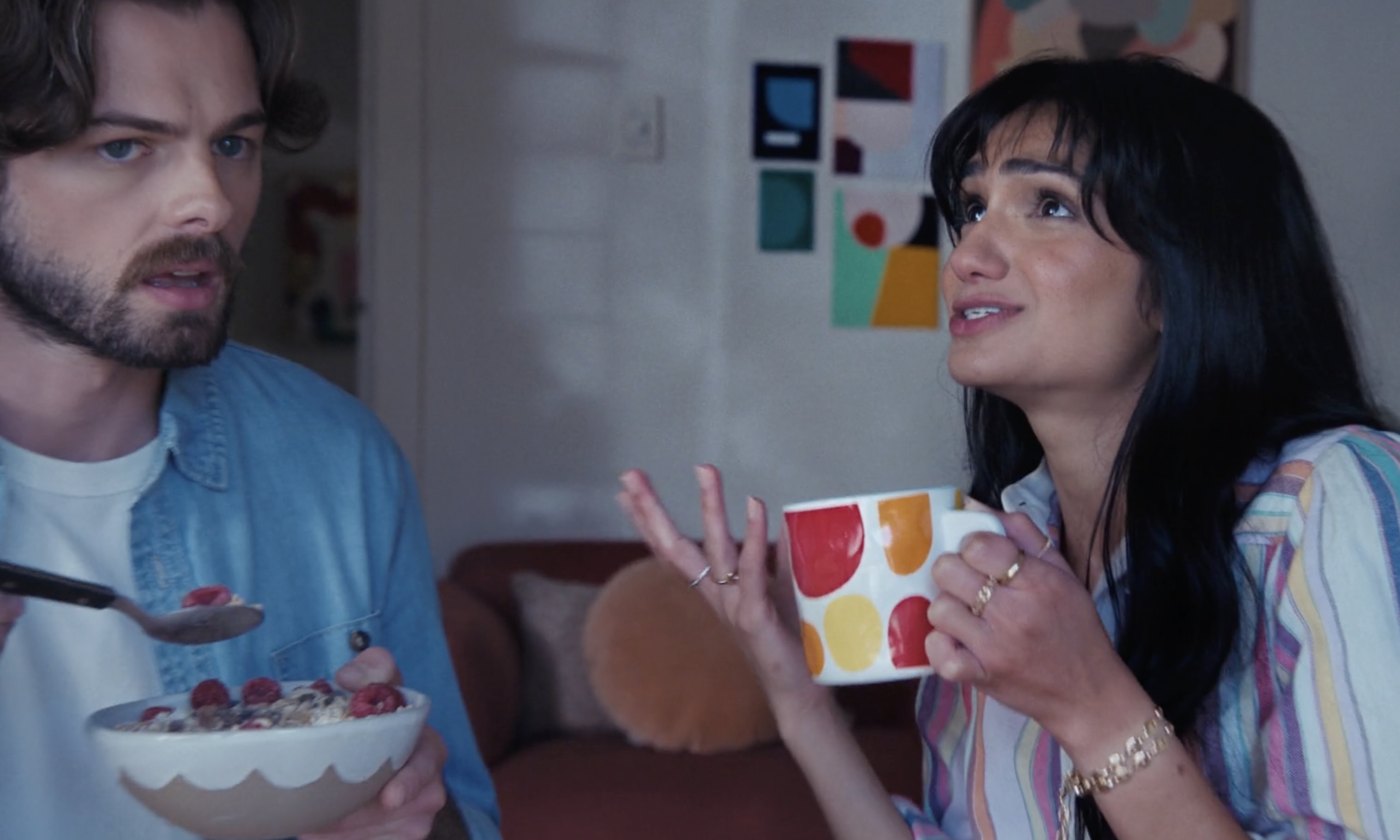Indoor shot of a man eating cereal and a woman drinking from a polka dot mug in a bright interior, creating a lifestyle scene.