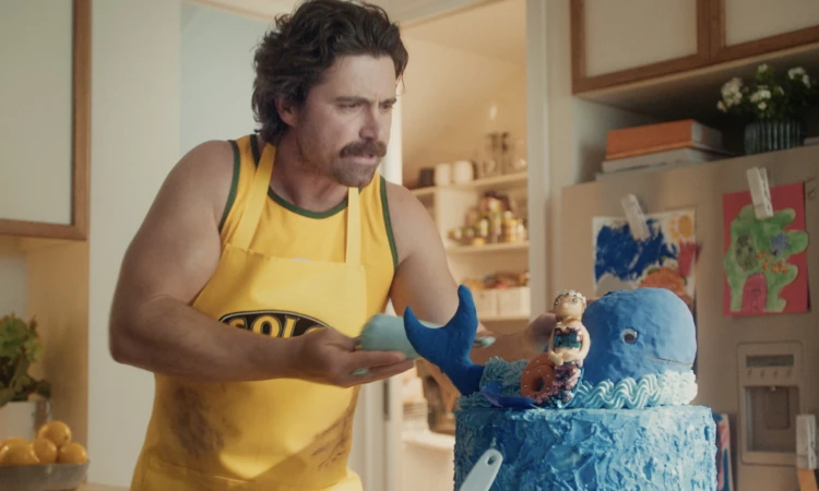 Man with a mustache in an apron decorates a whale-shaped cake in a bright kitchen with cabinets and playful children's artwork on the refrigerator.