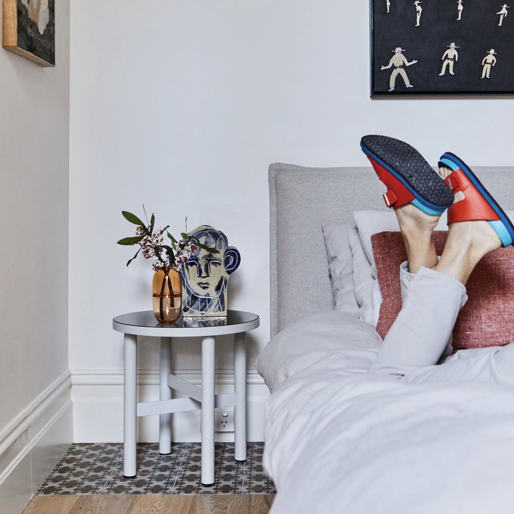 Interior photo of a bedroom featuring a person lying on a bed, with an end table and decorative items alongside, against a white wall.