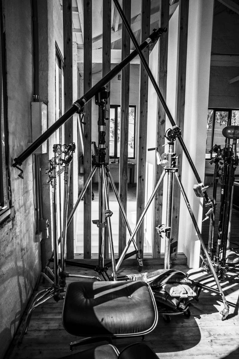 Monochrome shot of film studio equipment including tripods, boom arms, and a lounge chair, set against a textured wall and window.