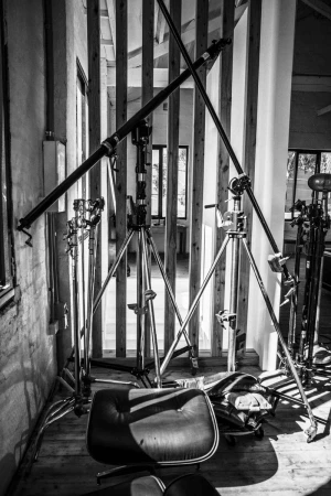 Monochrome shot of film studio equipment including tripods, boom arms, and a lounge chair, set against a textured wall and window.