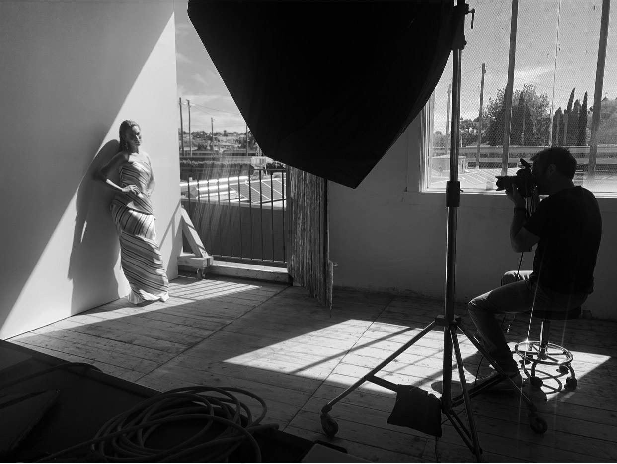 Black and white studio shot of a female model in a striped dress leaning against a wall, with a photographer taking her picture.