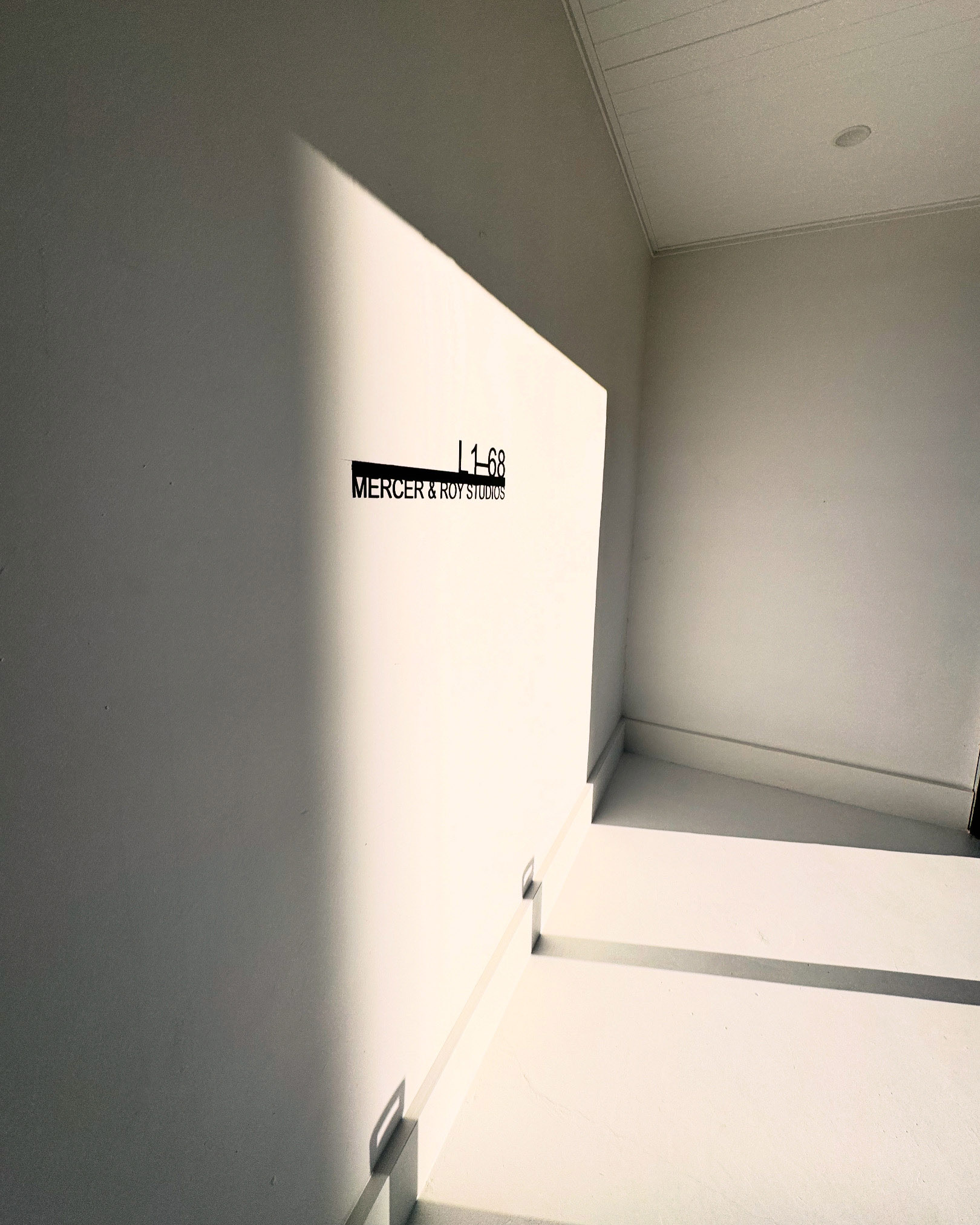 Interior architectural shot of a white hallway with sunlight and a black sign displaying 'Mercer & Koitshos' and 'L1-68'.