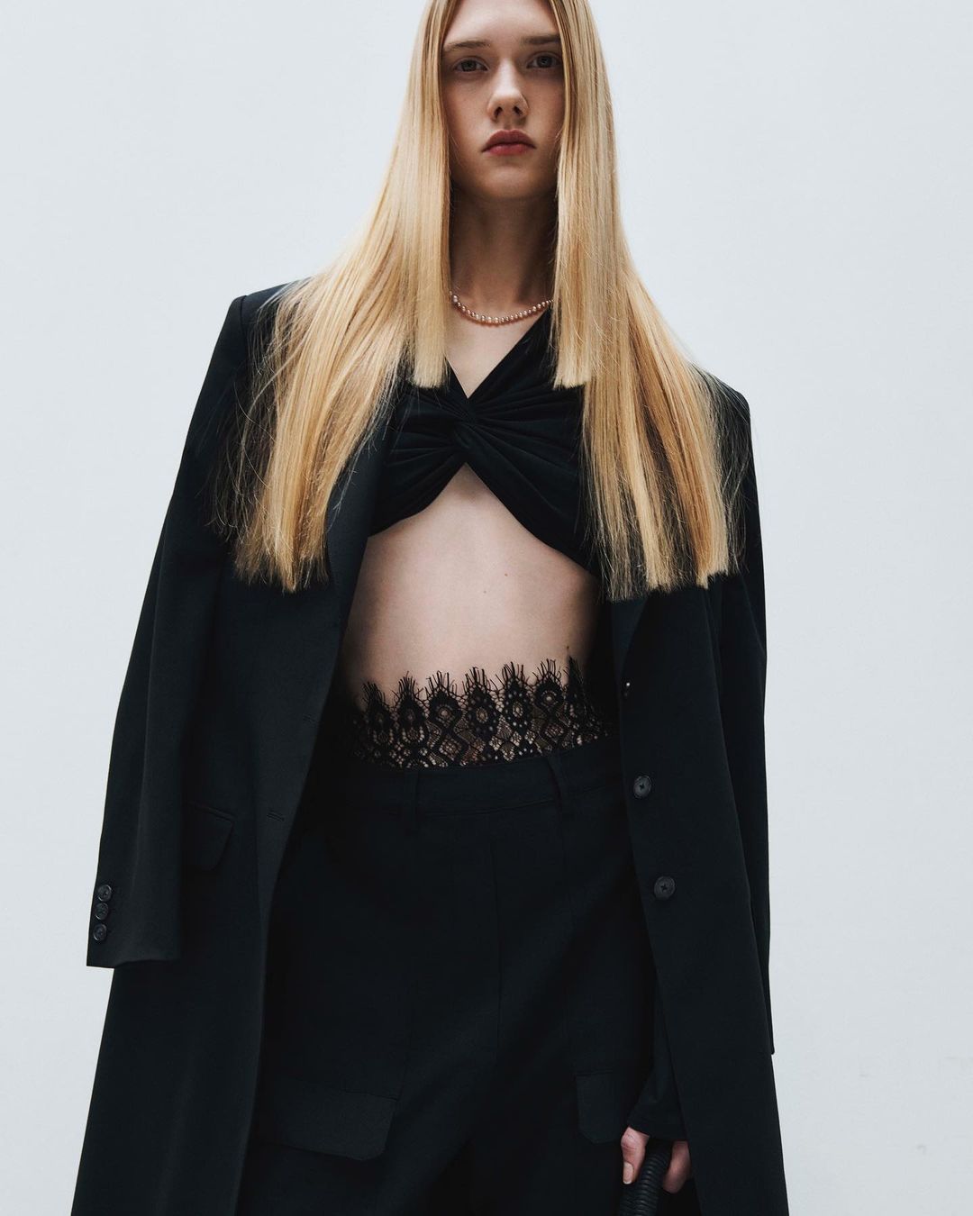 A slender young woman with long blonde hair poses in a black top, pants, and overcoat in front of a bright neutral backdrop.