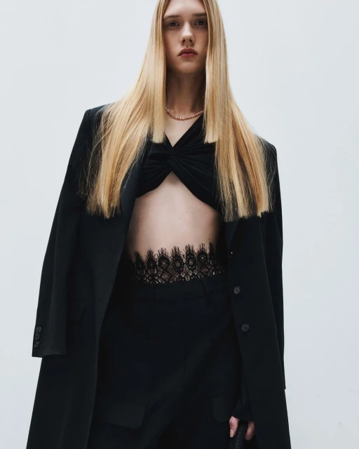 A slender young woman with long blonde hair poses in a black top, pants, and overcoat in front of a bright neutral backdrop.