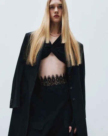 A slender young woman with long blonde hair poses in a black top, pants, and overcoat in front of a bright neutral backdrop.