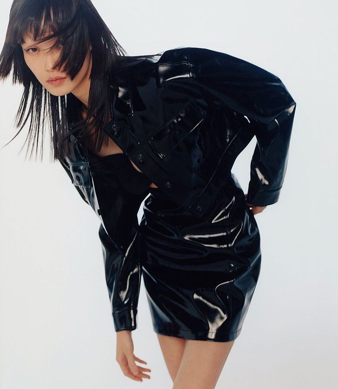 Portrait of Asian woman in shiny black jacket and mini-skirt on white background in fashion shot with her face partially obscured by hair.