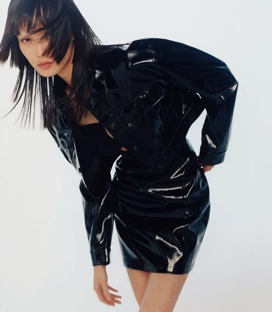 Portrait of Asian woman in shiny black jacket and mini-skirt on white background in fashion shot with her face partially obscured by hair.
