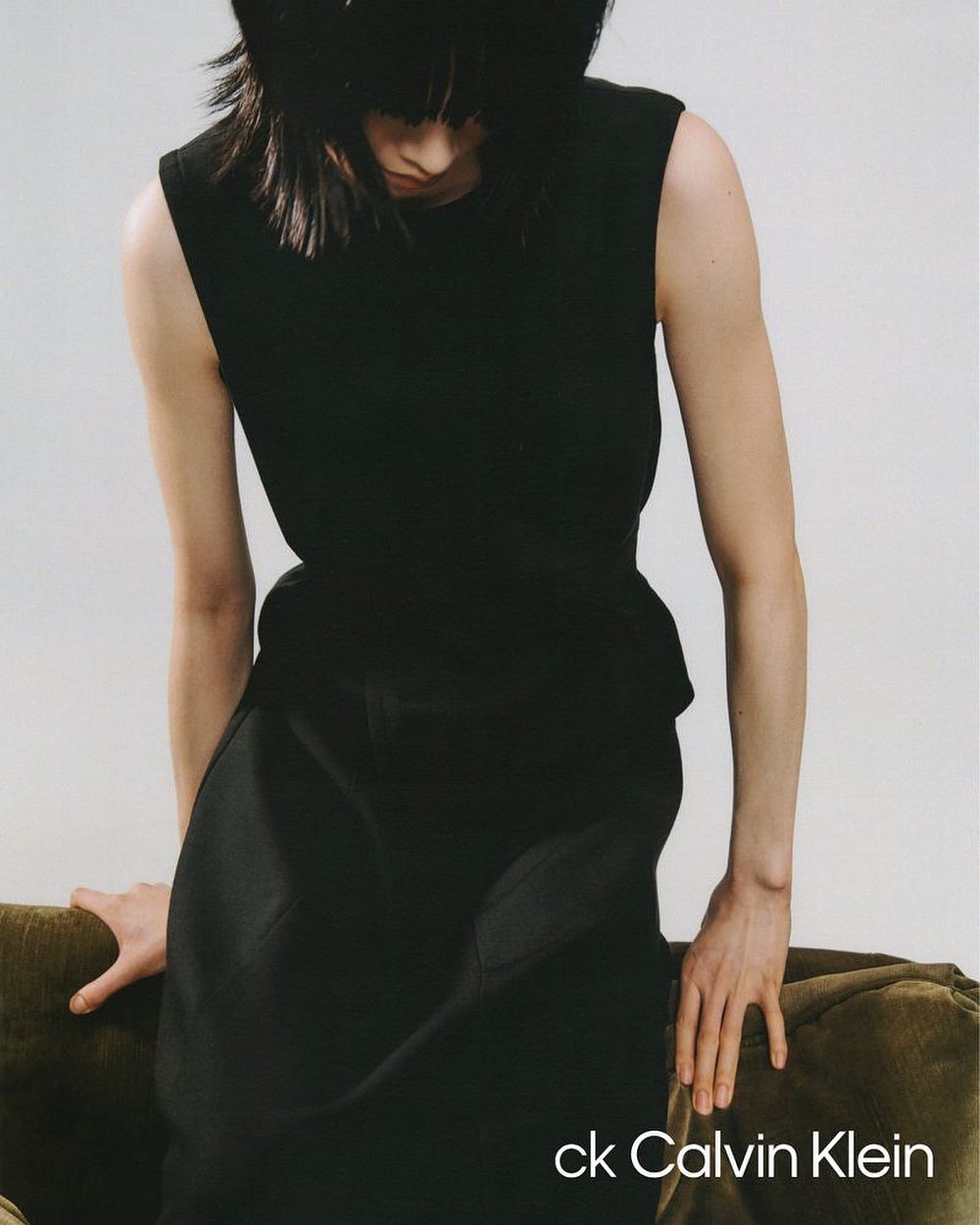 Fashion shot of an adult female wearing a black sleeveless top and skirt, posing indoors next to a green couch, with the ck Calvin Klein logo.