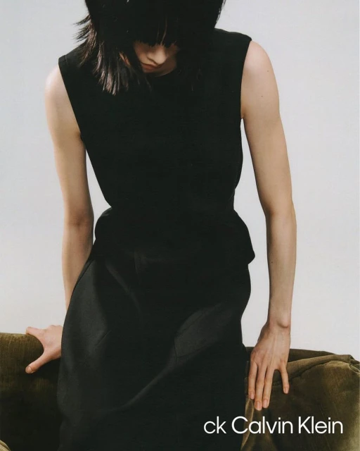 Fashion shot of an adult female wearing a black sleeveless top and skirt, posing indoors next to a green couch, with the ck Calvin Klein logo.