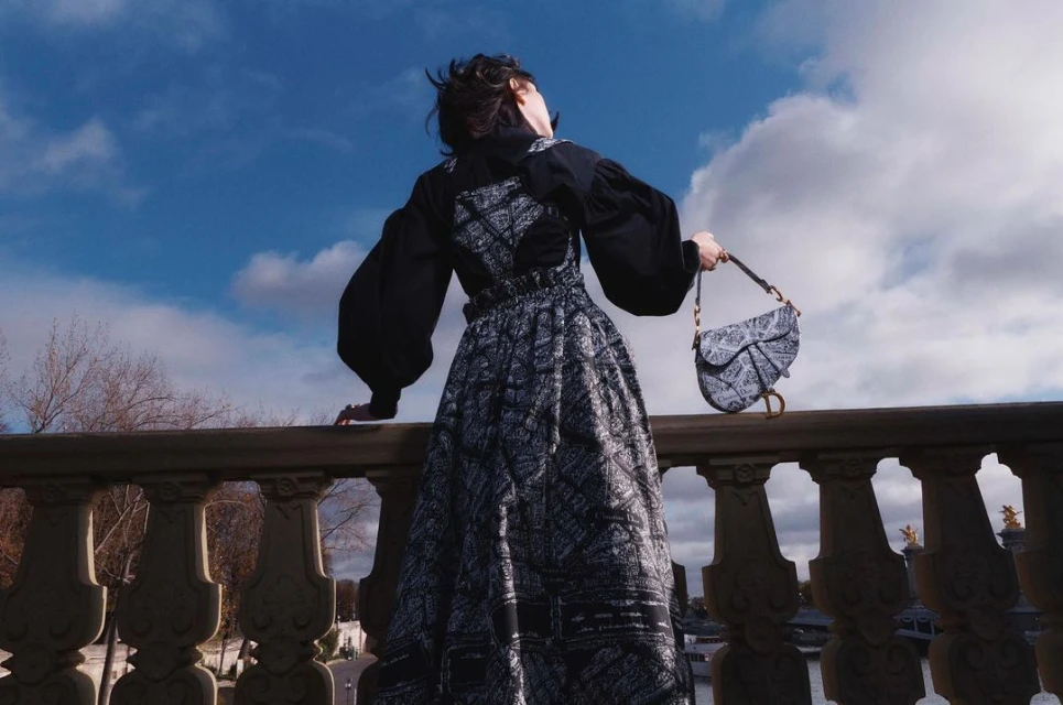 Woman stands on a stone bridge wearing a black and white dress, holding a Dior purse, with a blue sky and trees in the background.