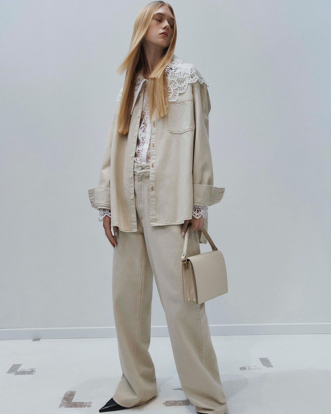 Adult female fashion model wearing beige denim outfit with lace collar and cuffs, holding a beige handbag in a photo studio.