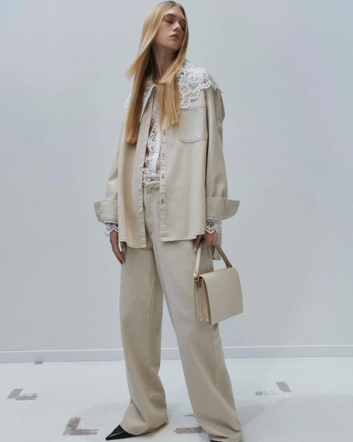 Adult female fashion model wearing beige denim outfit with lace collar and cuffs, holding a beige handbag in a photo studio.