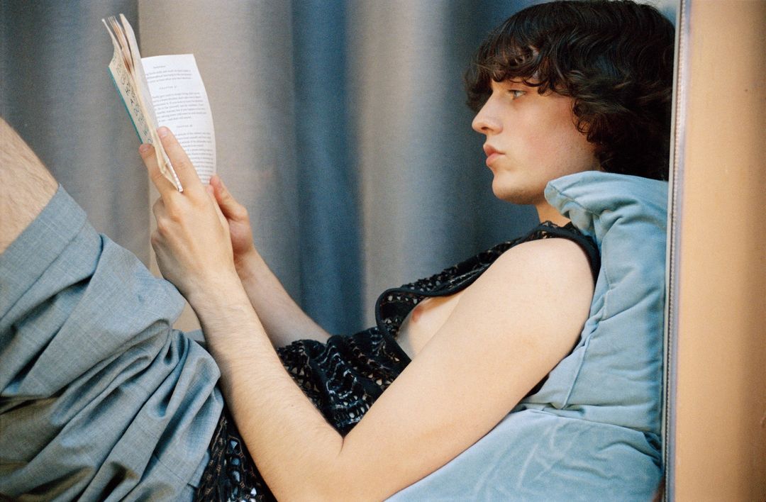 Young adult male reading a book indoors, resting against a light blue cushion and wearing grey pants and a dark, lacy shirt.