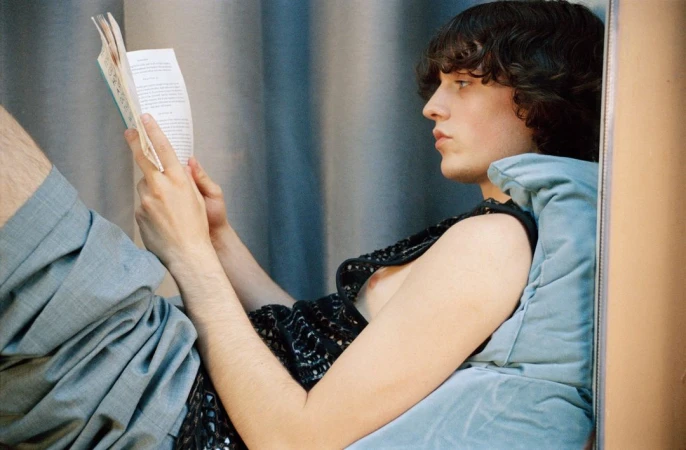 Young adult male reading a book indoors, resting against a light blue cushion and wearing grey pants and a dark, lacy shirt.